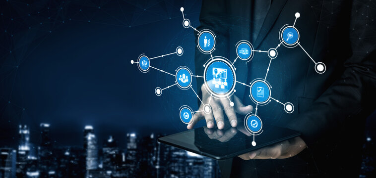 A businessman stands against a city skyline at night, interacting with a tablet that displays digital network icons, symbolizing technology and connectivity in business. Tessel - Powered by Adobe