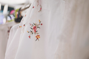 Beautifully embroidered white garments hang gracefully, showcasing intricate floral designs at a lively market in the morning sun, inviting all to admire their craftsmanship
