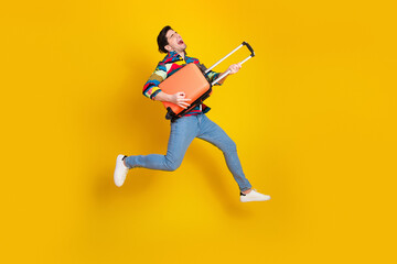 Joyful young man jumps with orange luggage against yellow background wearing colorful shirt jeans and white sneakers
