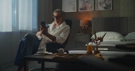 Stylish Senior Man in Soft White Bathrobe Uses Smartphone to Shop Online and Manage Payments. Calm...