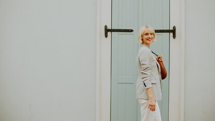 Smiling woman in a light gray blazer stands near a pastel blue door with a brown purse in hand on a sunny day