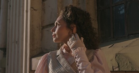Close up of a woman in historical clothing, looking pensively next to ancient stone columns, illuminated by soft golden evening light.
