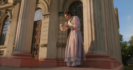 Naklejka premium An elegant woman in historical clothing stands between the massive, weathered columns of an ancient building and reads a letter in the warm evening light.