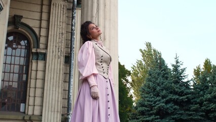 Fototapeta premium An elegant woman in a pastel historical dress stands next to a weathered column of an old building, with a dreamy, nostalgic expression on her face.