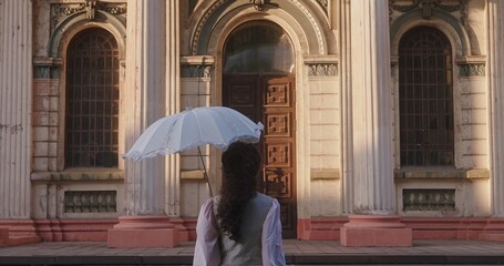 A woman in a historical dress holds a white umbrella and a suitcase, standing in front of majestic...