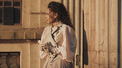 An elegant woman in historical clothing stands between the massive, weathered columns of an ancient building and reads a letter in the warm evening light.