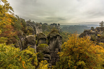 Misty view of the sandstone cliffs and forested valleys of Saxon Switzerland near the Bastei Bridge. Autumn colors blend with fog and soft light, creating a mysterious and atmospheric mountain scene.