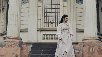 Fototapeta premium An elegant woman in a pastel historical dress stands next to a weathered column of an old building, with a dreamy, nostalgic expression on her face.
