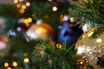 Close-up of Christmas tree branches adorned with decorations. Prominent features include a large, glossy golden ornament embellished with sequins, creating a sparkling texture.