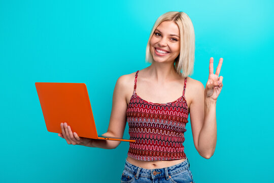 Joyful young woman with orange laptop poses against teal background and flashes peace sign in stylish casual top during colorful lifestyle and advertising vibe