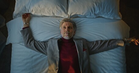 Fototapeta premium Aged Man in Suit Happily Falls Down on White Bed in Cozy Hotel Room. Tired From Traveling on Business Trip, Man Rests on Bed. Concept of Life in Hotel, Traveler Fatigue, Business Trip. Top View.