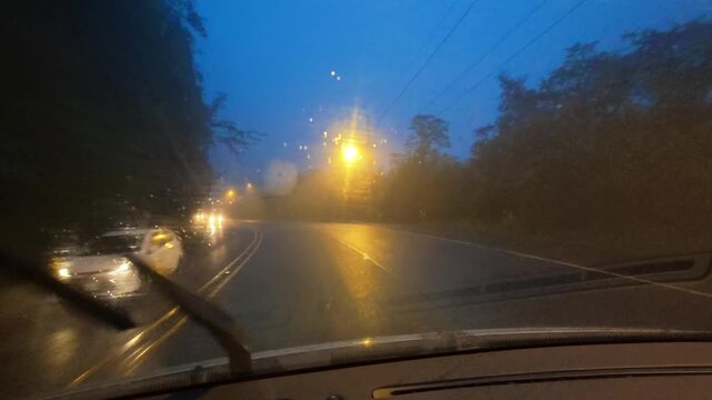 View from inside a moving car driving through a rainy road at dusk or dawn. The windshield wipers are active as water blurs the view.