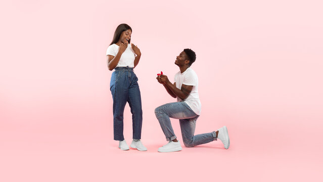 A joyful black man kneels and presents an engagement ring to his thrilled girlfriend, asking her to marry him. They are in front of a soft pink background, capturing a beautiful moment of love.