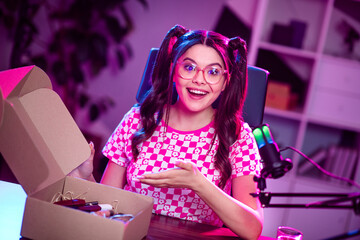 Smiling young female blogger presenting unboxing content in her neon-lit workspace during an online...