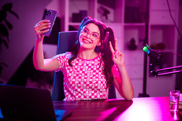 Charming female influencer live-streaming with microphone and phone under neon lights, expressing creativity and enthusiasm