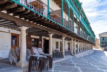 A view within Chinchón's Plaza Mayor, highlighting the distinctive three-story arcaded buildings with turquoise wooden balconies and heavy stone columns in the foreground.
