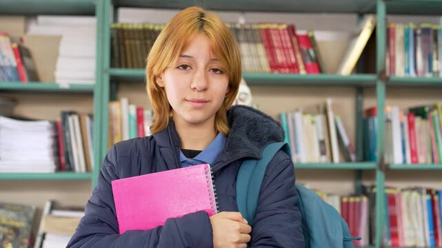 Portrait of a smiling female high school student holding a pink notebook in the school library