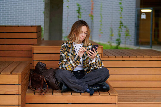 woman using smartphone on wooden steps outdoors. casual fashion style with plaid shirt and black boots. urban environment and technology use in modern lifestyle setting - Powered by Adobe