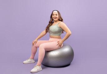 Cheerful plus size European millennial woman in stylish sportswear sits on a fitness ball against a violet background. This setting promotes body positivity and exercise during lockdown.