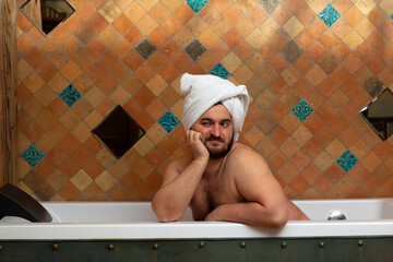 Horizontal photo. Fat naked man with beard, bare shoulders, white towel on head sits in bathtub in hotel bath room. Concept of hygiene, wash, skin care, relax, father day, 8 march, rest, spa, purity