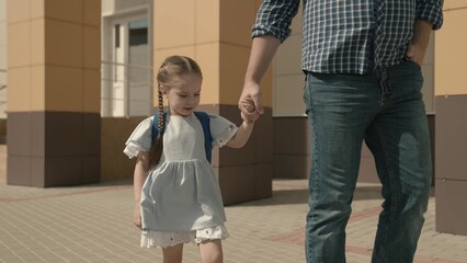 father holds happy daughter by the hand, accompany little child with backpack to school, study in preschool preparation, walk through schoolyard, first grade student with school bag on his shoulders.