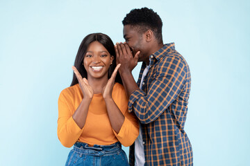 Naklejka premium A young black man whispers a secret into the ear of an excited woman with a bright smile. They are enjoying a playful moment together against a soft blue and purple background.