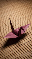 Traditional Japanese origami crane in a rich burgundy color resting on a bamboo mat. A serene still life representing peace, hope, and the delicate art of paper folding, with dramatic lighting.
