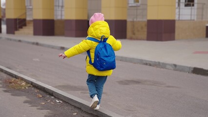 a little child goes to school with blue backpack, funny school weekdays kid, girl student walks around the schoolyard of the school building, preschool baby with bag on his shoulders, happy childhood.