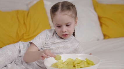 Little girl eats potato chips from a plate lying on the bed in the bedroom, delicious food with spices and flavors for kid pleasure, crispy baby snack indoors, child enjoying the taste of potatoes.