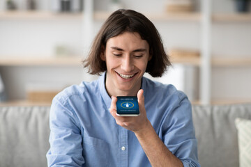 A young man smiles while communicating with a voice assistant device in a stylish living room. Natural light fills the space, creating a cheerful atmosphere.