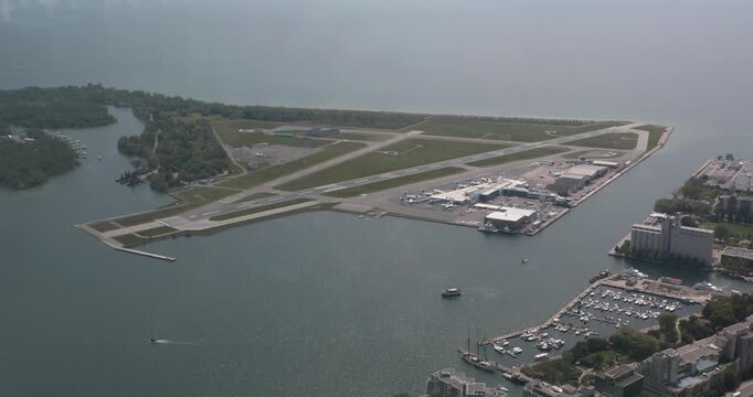 Aerial shot of Toronto's island airport on Lake Ontario. Runways, terminal, small planes, ferry channel and surrounding marinas sit on a narrow strip of land. Calm water, soft haze, urban proximity.