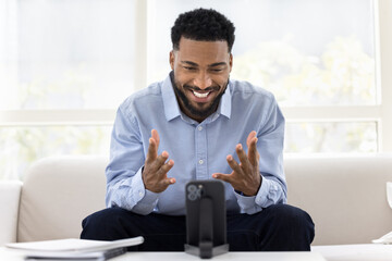 African man sits on sofa, gesturing while interacting on smartphone
