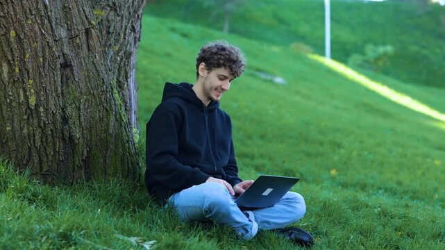 Happy young man sitting near the tree in the park while working with laptop and typing. Autumn season. Education, working concept. Slow motion