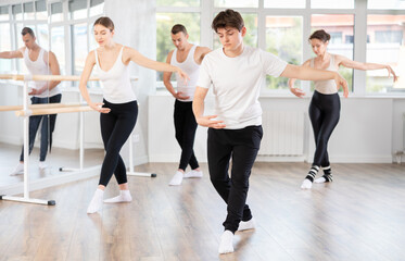 Fototapeta premium Happy young guy in sportswear confidently leading ballet class with dancers following movements in modern dance studio
