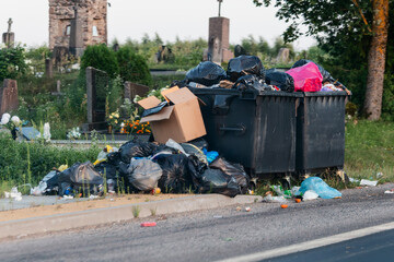 An overflowing dumpster and a large pile of trash bags are discarded on the side of a road next to an old cemetery. Lithuania.
