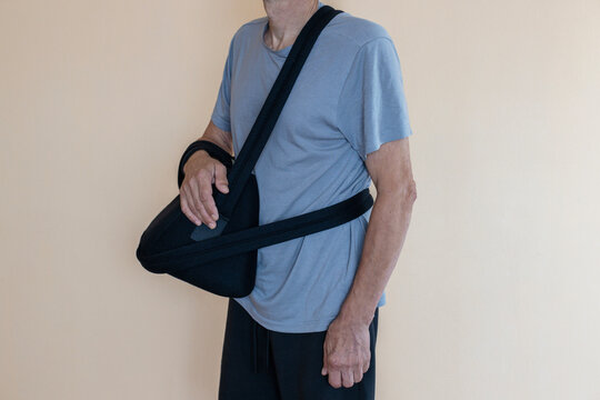 An older adult man stands wearing a black arm sling to support his injured arm against a plain, light-colored wall.