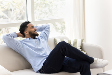 Carefree African guy relaxing on couch in well-lit living room
