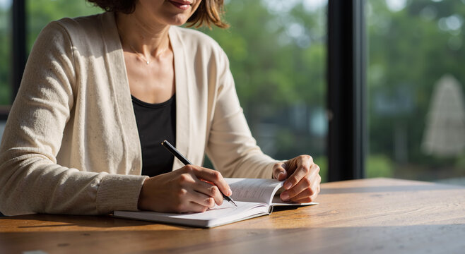 woman writing in journal at wooden table by large window on a sunny day