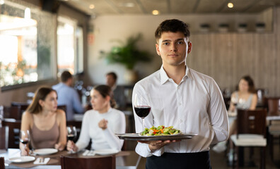 Young male waiter in uniform posing with tray and glass of red wine in his hands in restaurant