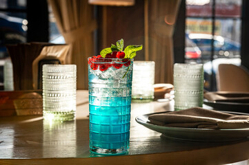 Blue cocktail with mint and berries on restaurant table. Blue cocktail with mint and red currant in glass with ice on table in stylish restaurant setting with sunlight.