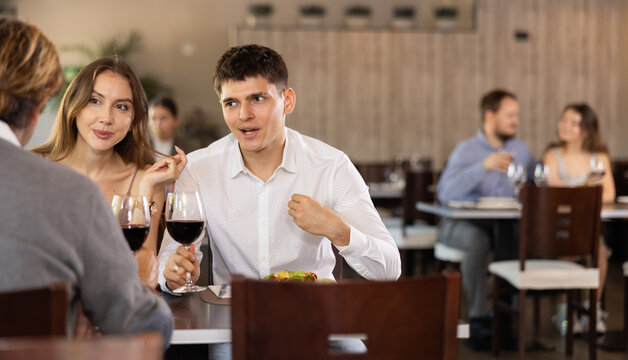 Couple young man and woman with adult man eat drink wine and chat in restaurant