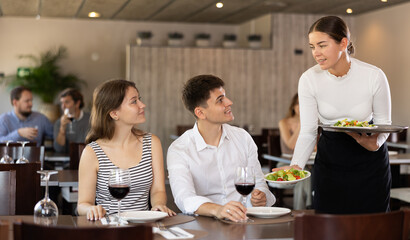 Young woman waitress brings order from couple young man and young woman in restaurant