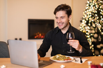A man sits at a table with a plate of pasta and a glass of wine, smiling at his laptop, surrounded by cozy winter decorations and a festive tree, enjoying his evening.