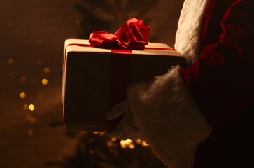 A festive scene featuring a Santa Claus figure holding a wrapped Christmas gift with a red ribbon in front of a beautifully lit Christmas tree.
