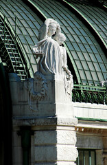 A sculpture of two figures, possibly angels, on a stone pedestal, with a green metal roof in the background.