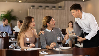 Couple resting, sitting in restaurant and ordering food. Waiter guy tells couple of cafe about dish from chef