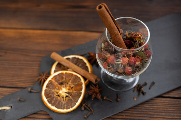 A glass glass with spices, a stick of cinnamon, star anise and cloves with dried hawthorn. Slices of dried orange are laid out on black slate.