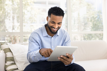 Happy young African man using modern tablet seated on sofa