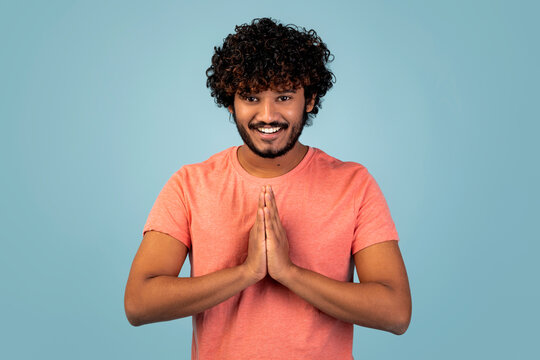 A happy young Indian man stands against a blue studio background, smiling broadly while holding his hands in a namaste gesture to express gratitude and thankfulness.