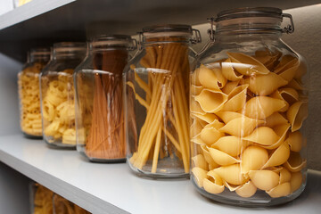 Row of glass jars filled with assorted dry pasta shapes on a shelf, showcasing pantry staples in clear storage with shallow depth of field.